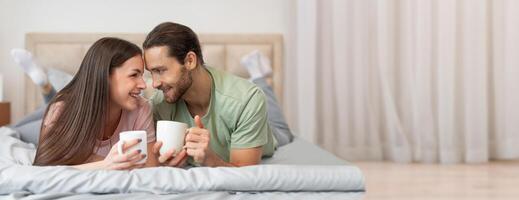 A couple lies comfortably on their bed, smiling and sharing conversations over steaming cups of coffee. The bright sunlight filters through sheer curtains, creating a warm atmosphere. photo