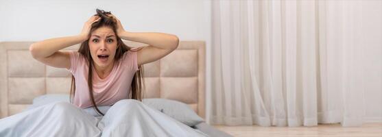 A woman sits up on her bed, visibly distressed, with her hands on her head. The soft lighting highlights her anxious expression in a calm bedroom setting. photo
