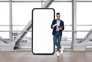 Arab man in a business suit stands against a large blank digital display at an airport terminal, looking at his tickets while holding a smartphone and a document, waiting for his flight. photo