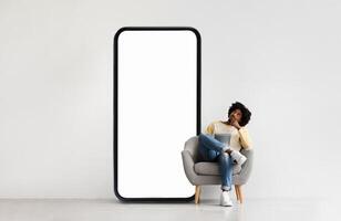 A person relaxes in a modern chair while contemplating, situated beside a giant smartphone-shaped screen. The indoor setting has a minimalist design and neutral tones. photo