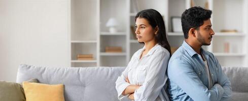Indian couple faces away from each other in a contemporary living room, each appearing distant and upset, reflecting a moment of emotional disconnect during a disagreement. photo