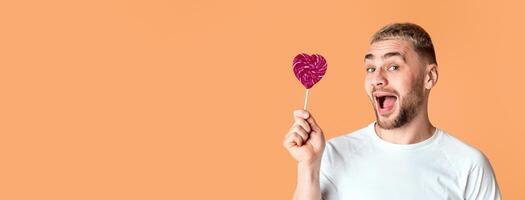 Emotional man with open mouth holds heart lollipop, panorama, free space photo