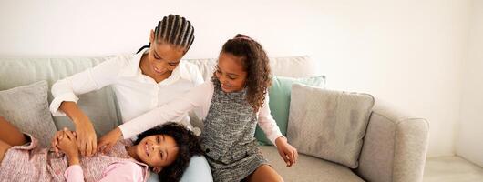African American mother sits on the couch as her two daughters playfully interact, showcasing laughter and love in a cozy living room setting during the afternoon. photo