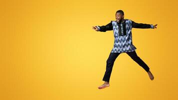 Excited black man in colorful authentic costume running towards copy space for advertisement, pink studio background. Shocked african american guy jumping and pointing at empty space, full length shot photo