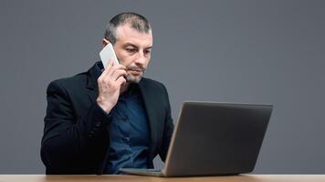 Distant Work. Mature Entrepreneur Talking On Cellphone Working On Laptop Sitting On Gray Studio Background photo