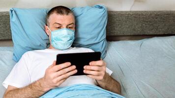 Coronavirus Quarantine. Middle-Aged Man In Mask Using Digital Tablet Watching Movie Lying In Bed At Home. Selective Focus photo
