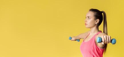 Young Sporty Woman Training With Dumbbells, Standing Over Yellow Background In Studio, Panorama With Free Space photo