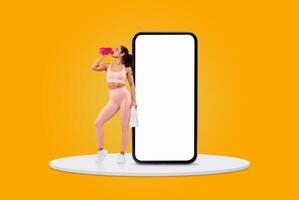 A woman dressed in pink athletic clothing stands confidently, sipping water from a pink bottle beside a large blank smartphone on a vibrant yellow background. photo
