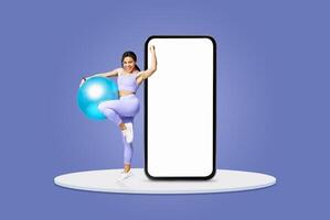 A young woman in purple activewear enthusiastically holds a fitness ball while playfully leaning against a large, blank phone display. The vibrant background enhances the energy of the moment. photo
