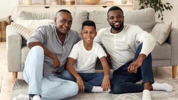 Family Portrait. Happy Black Boy With Father And Grandfather Looking At Camera While Sitting On Floor In Living Room photo