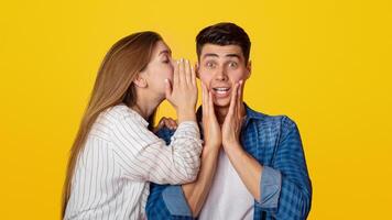Omg, shocking gossip, secrets and wow. Excited millennial lady tells in ear to surprised guy something amazing, funny man does not believe, presses his hands to cheeks, isolated on yellow background photo
