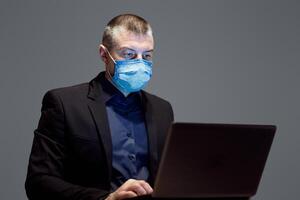 Businessman In Protective Medical Mask Working On Laptop Sitting Over Gray Studio Background. Work From Home. Selective Focus photo