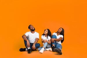 Free Place For Your Advert. Smiling African American Family Of Three People Sitting On The Floor, Looking Up At Empty Copy Space Isolated On Yellow Studio Background, Vertical Shot, Banner photo