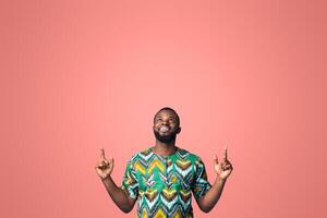 Happy black guy in african costume pointing up at free space, showing advertisement on blue studio background. Positive african american man looking up, directing at empty space for text photo