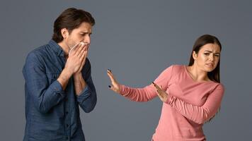 Young woman feeling disgust next to her sick male friend, trying to keep distance, standing together on grey background in studio, free space photo