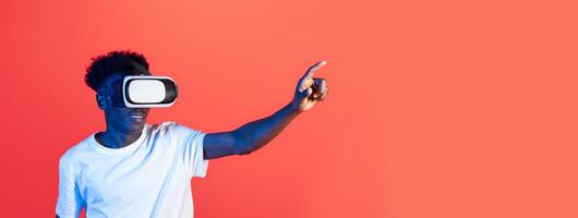 African American young man is fully engaged with a virtual reality headset, pointing as if navigating through an immersive digital experience in a brightly colored studio. photo