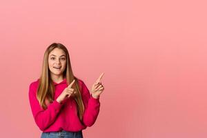 Funny smiling teenager girl with open mouth points with fingers towards an empty space, on pink background, studio shot photo
