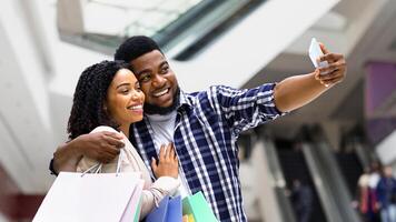 Shopping Fun. Joyful black couple taking selfie in mall center, using smartphone, holding lots of paper bags photo
