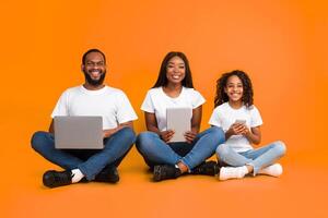Gadget Generation. Happy African American family of three people holding different electronic devices in hands, sitting in a row on the floor over yellow studio background, posing looking at camera photo