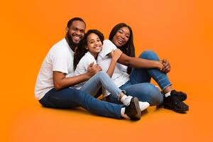 Portrait of happy smiling three black people hugging, having fun spending time together, dad embracing family, looking at camera, sitting on the floor isolated on yellow orange studio background photo