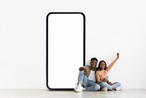 A cheerful black couple sits on the floor, relaxed and smiling. They lean against a large blank phone screen, enjoying their time together and engaging in conversation. photo