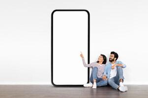 A young Indian couple sits on the floor, smiling and pointing at a large blank smartphone display. They are in a modern room with minimal decor, creating a relaxed atmosphere. photo