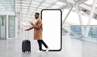 A traveler black guy stands in an airport terminal, pulling a suitcase while looking at his phone. A large blank screen looms in the background, adding to the contemporary atmosphere. photo
