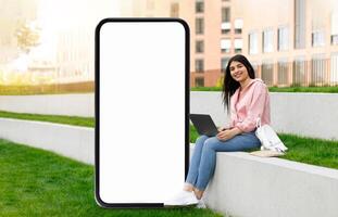 A young woman sits on a grassy area in a park, focused on her laptop while a large blank smartphone display stands beside her. Sunlight bathes the scene, creating a relaxed atmosphere. photo
