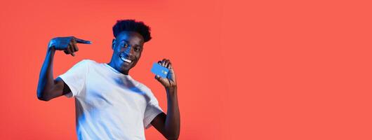 Black man is standing in front of a red backdrop, smiling and holding a credit card in his right hand. He is wearing a white t-shirt and pointing to himself with his left hand. photo