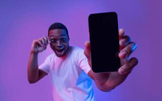 Cool Mobile App. Mockup Image Of Smartphone With Black Blank Screen In Hand Of Excited African American Guy In Sunglasses Standing In Vivid Neon Light Over Purple Studio Background, Copy Space photo