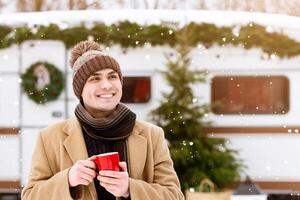 A man wearing a warm coat and knitted hat smiles as he sips a hot beverage while standing in the snow. The backdrop features a decorated trailer, enhancing the winter atmosphere, copy space photo