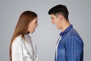 Breakup of relations, quarrel, problems in couple and scandal. Angry aggressive young man and lady yelling at each other with open mouths, isolated on gray background, studio shot, profile, free space photo
