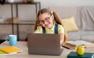 Distance Education. Smiling female pupil in eyeglasses sitting at desk, using laptop and typing on keyboard, doing homework, watching tutorial, lecture or webinar, studying online at home. E-learning photo