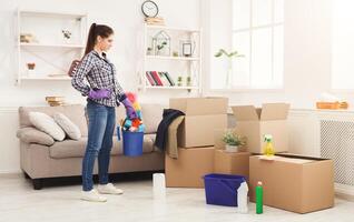 New home cleaning. Young woman tiding up after moving to new appartment. Girl with various detergents, rags and mops in living room full of carton boxes. Household and spring-cleanig concept photo