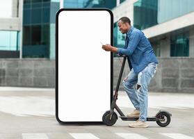A young black man rides an electric scooter while checking his phone, positioned next to a large blank display screen in an urban area during daylight hours. photo