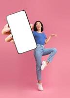 Check out this new mobile application. Excited young Asian lady jumping, shouting OMG, presenting smartphone with empty screen over pink studio background, mockup for website or app photo