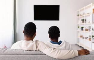 Rest And Relax. Rear back view of afro man and woman sitting on the sofa in living room, watching television show together, looking at blank plasma tv on the wall with isolated empty monitor, mock up photo