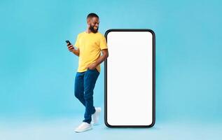 Cheery millennial black guy with mobile device looking at giant smartphone on blue background, mockup for mobile app on white screen. Cellphone website template, cool online store ad photo