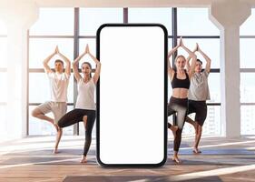 A group of four individuals performs yoga poses in a bright, modern studio. The large windows let in natural light, and a smartphone stands in the center, inviting a digital connection to wellness. photo