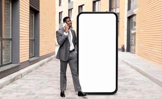 A well-dressed black businessman stands outside, talking on his smartphone while leaning against a large, blank billboard. The urban setting features modern buildings and a clear sky. photo