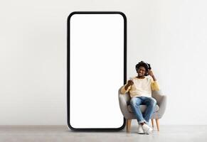 A young black man sits comfortably in a chair, enjoying his smartphone as he listens to music. Next to him stands a large blank screen, creating a minimalist atmosphere. photo