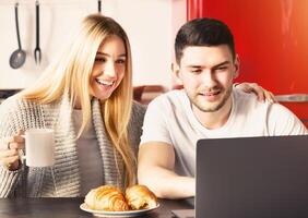 Lovely family of two having breakfast while using laptop in kitchen, panorama photo