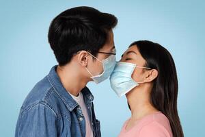 Romantic Young Chinese Couple Kissing In Protective Medical Face Masks Against Virus Spread, Standing Over Light Studio Background, Free Space photo