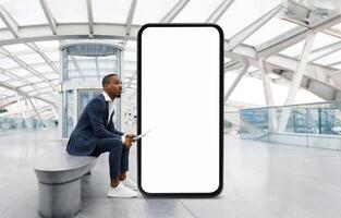 African American man in a suit is seated on a bench in a contemporary architectural space, appearing thoughtful as he waits next to a large, empty digital display. photo