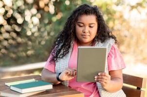 Hispanic lady student using digital tablet websurfing sitting at table in park. Young woman with computer learning and reading online books outside. Learner preparing for college exam photo