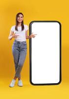 Mobile App Advertisement. Full Body Length Of Happy Woman Showing Pointing At Big Huge White Empty Smartphone Screen Standing Over Orange Studio Background. Check This Out, Cellphone Display Mock Up photo