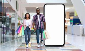 A joyful black couple strolls through a modern shopping mall, holding colorful shopping bags. They walk beside a large, empty display screen, creating a vibrant atmosphere. photo