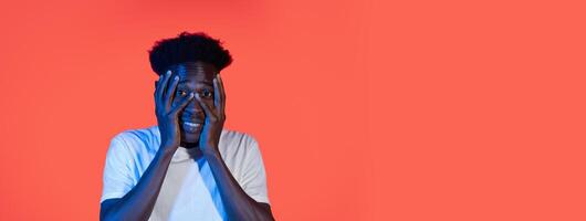 A young black man stands in front of a bright orange backdrop, showcasing a surprised expression with hands on his face. His casual attire contrasts with the vivid color around him. photo