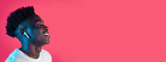 A young black man enjoys music through wireless earbuds, smiling with his eyes closed. The vibrant pink backdrop enhances the joyful atmosphere, capturing a moment of relaxation and enjoyment. photo