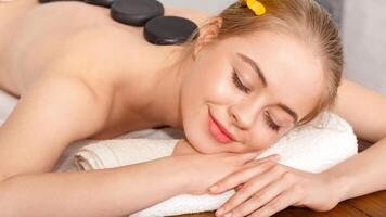 Health and beauty concept. Smiling woman with closed eyes with hot stones on back lie on white towel and on massage table photo